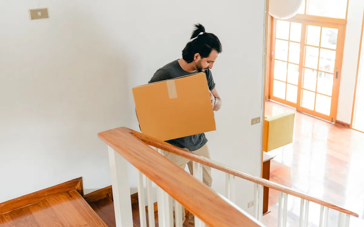 Eine Person trägt einen Karton durch eine Wohnung mit Treppenaufgang